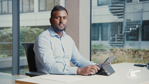 HZD-Mitarbeiter sitzt mit Laptop am Schreibtisch und schaut in die Kamera