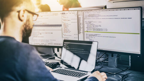 Blick von schräg hinten auf Mann am PC-Arbeitsplatz mit Monitoren, die Programmiercodes zeigen