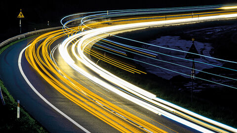 Straßenkurve mit Baustellenschild bei Nacht, die in Langzeitbelichtung fotografier wurde und die Lichter der fahrenden Autos als gelbe und weiße Linien zeigt