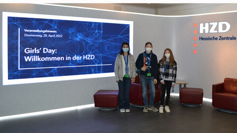 Man sieht die LED-Wand mit der Textanzeige "Veranstaltungshinweis 28. April 2022 Girls'Day Willkommen in der HZD". Rechts daneben stehen drei der vier Mädchen, die die HZD besucht haben.
