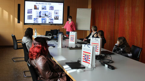 Die Schülerinnen sitzen mit viel Abstand und Maske an den Seiten eines Konferenztischs und schauen auf einen Bildschirm. Die Gleichstellungsbeauftragten Dr. Anette Knedla erläutert, für welche Kunden die HZD arbeitet und welche Aufgaben sie dabei übernimmt. Auf dem Tisch stehen Taschen mit dem Hessen-Wappen.