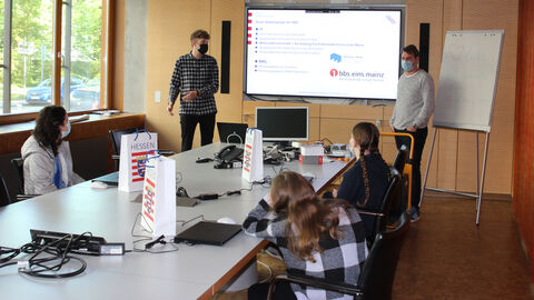 Man sieht drei der Girls'Day-Besucherinnen, die am Konferenztisch sitzen und nach vorne schauen. Neben einem großen Bildschirm in einer Holzwand stehen zwei jungen Männer, die anhand einer Powerpoint-Präsentation die Phasen eines dualen Studiums erläutern.
