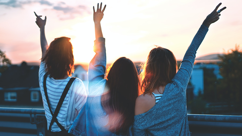 Drei junge Mädchen von hinten, die mit nach oben ausgesteckten Armen den Sonnenuntergang betrachten