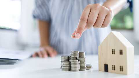 Tisch mit Spielzeughaus, neben dem eine weibliche Hand Münzen stapelt