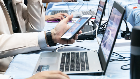 Close-up auf Tischreihe mit offenen Laptops und angedockten Smartphones, an denen Lernende arbeiten