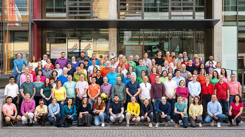 Gruppenfoto mit über 100 bunt gekleideten Mitarbeiterinnen und Mitarbeitern vor dem Neubau der HZD in Wiesbaden