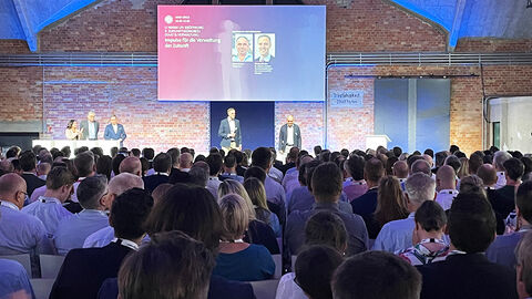 Blick über das Publikum hinweg auf die Vortragsbühne des Zukunftskongresses, wo Staatssekretär Dr. Markus Richter vom BMI die Keynote zur Veranstaltung gibt