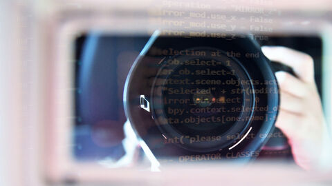 Großaufnahme von Kameralinse mit unscharf gehaltenen Hintergrund, in dem eine Hand am Kameraauslöser zu erkennen ist; darüber gelegt eine Grafik aus Programmierbefehlen