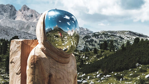 Hochgebirgslandschaft mit bewölktem Himmel und Skulptur eines Astronauten, in dessen Helmvisier sich die Umgebung spiegelt