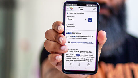Mann mit Handy in der weit vorgestreckten Hand; auf dem Display ist die Benutzeroberfläche der von der HZD entwickelten HessenApp zu sehen