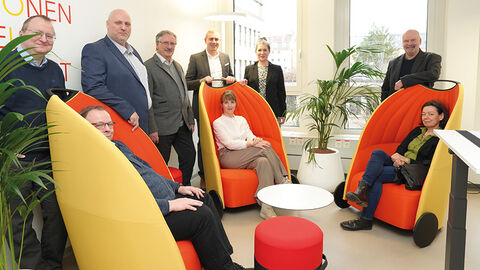 Beschäftigte der HZD - darunter der Technische Direktor Thomas Kaspar - stehen hinter drei gelb-roten Akustiksesseln, in denen Personen Platz genommen haben.