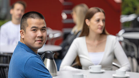Junger Mann, der im Innenhof der HZD in einer Gesprächsrunde am Tisch sitzt und über die Schulter in die Kamera blickt