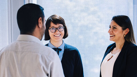 Drei lachende Menschen beim Gespräch vor einem deckenhohen Fenster in einem Büro der HZD
