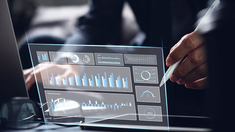 Close-up auf Schreibtisch, auf den das Hologramm eines Monitors mit unterschiedlichen Grafiken zur Leistungsdarstellung projiziert ist