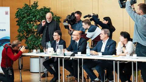 Justizminister Heinz am Tisch mit HZD-Vertretenden und umringt von Journalisten