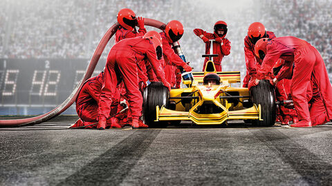 Gelber Rennwagen beim Boxenstopp an einer Rennbahn, der von einer großen Crew in roten Overalls gewartet wird