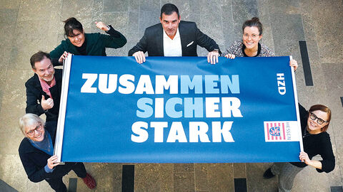 Blick ins Atrium der HZD, in dem sechs Personen ein Banner mit dem Jahresmotto „Zusammen sicher stark“ hochhalten 