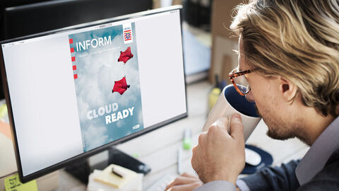Ein blonder Mann mit Brille an einem Schreibtisch sitzend, der gerade eine Tasse zum Mund führt, blickt auf den Computerbildschirm vor sich, auf dem das Titelbild einer INFORM-Ausgabe zu sehen ist
