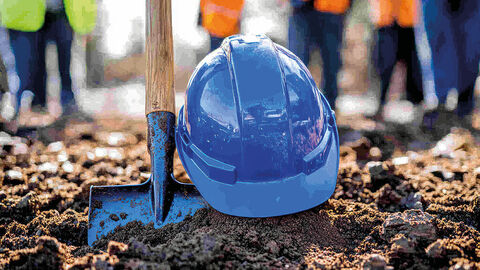 Close-up auf einen in der Erde steckenden blauen Spaten, daneben ein Schutzhelm in demselben Blau